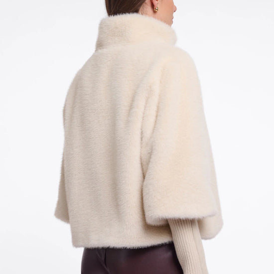 A woman shown from the back wearing a cream-colored, fluffy cropped cape jacket over a ribbed cream sweater and dark brown pants, against a plain white studio background.
