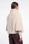 A woman shown from the back wearing a cream-colored, fluffy cropped cape jacket over a ribbed cream sweater and dark brown pants, against a plain white studio background.