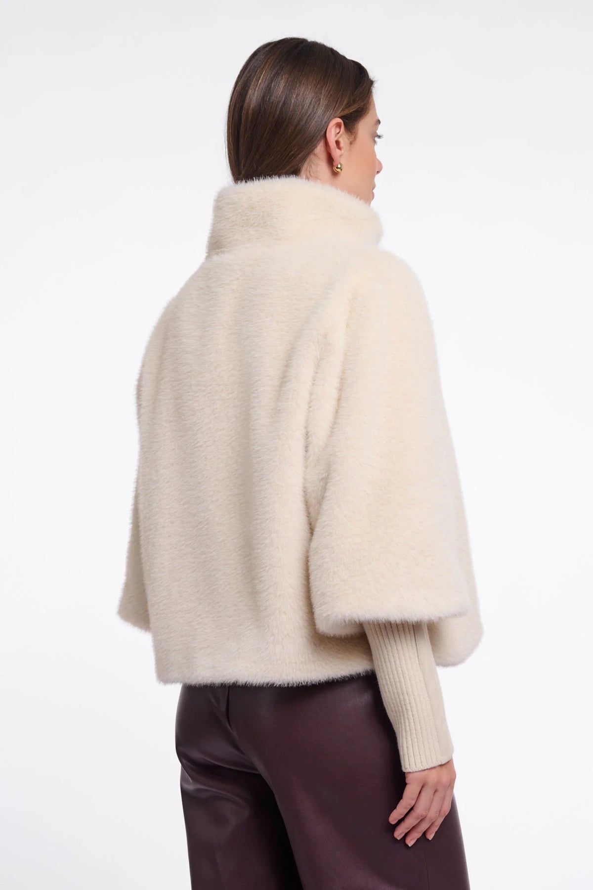 A woman shown from the back wearing a cream-colored, fluffy cropped cape jacket over a ribbed cream sweater and dark brown pants, against a plain white studio background.
