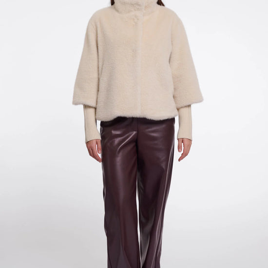 A model stands against a light gray studio background wearing a cream-colored fuzzy mock-neck jacket over a light beige top, paired with dark brown wide-leg faux-leather trousers and beige pointed-toe heels.