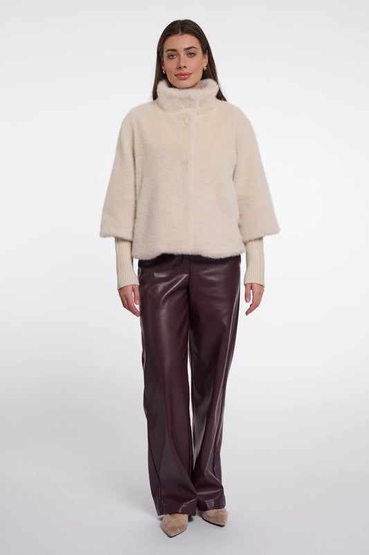 A model stands against a light gray studio background wearing a cream-colored fuzzy mock-neck jacket over a light beige top, paired with dark brown wide-leg faux-leather trousers and beige pointed-toe heels.