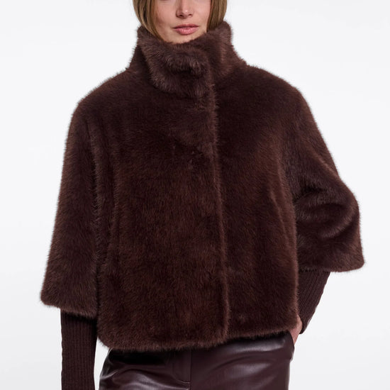 A model wearing a dark brown plush faux fur jacket with a high collar and wide sleeves, paired with dark brown faux leather pants, against a plain white background.