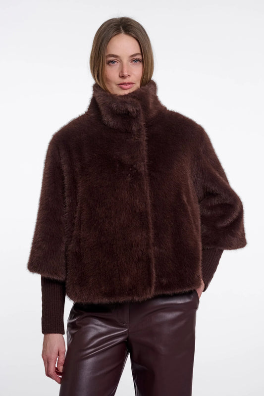 A model wearing a dark brown plush faux fur jacket with a high collar and wide sleeves, paired with dark brown faux leather pants, against a plain white background.