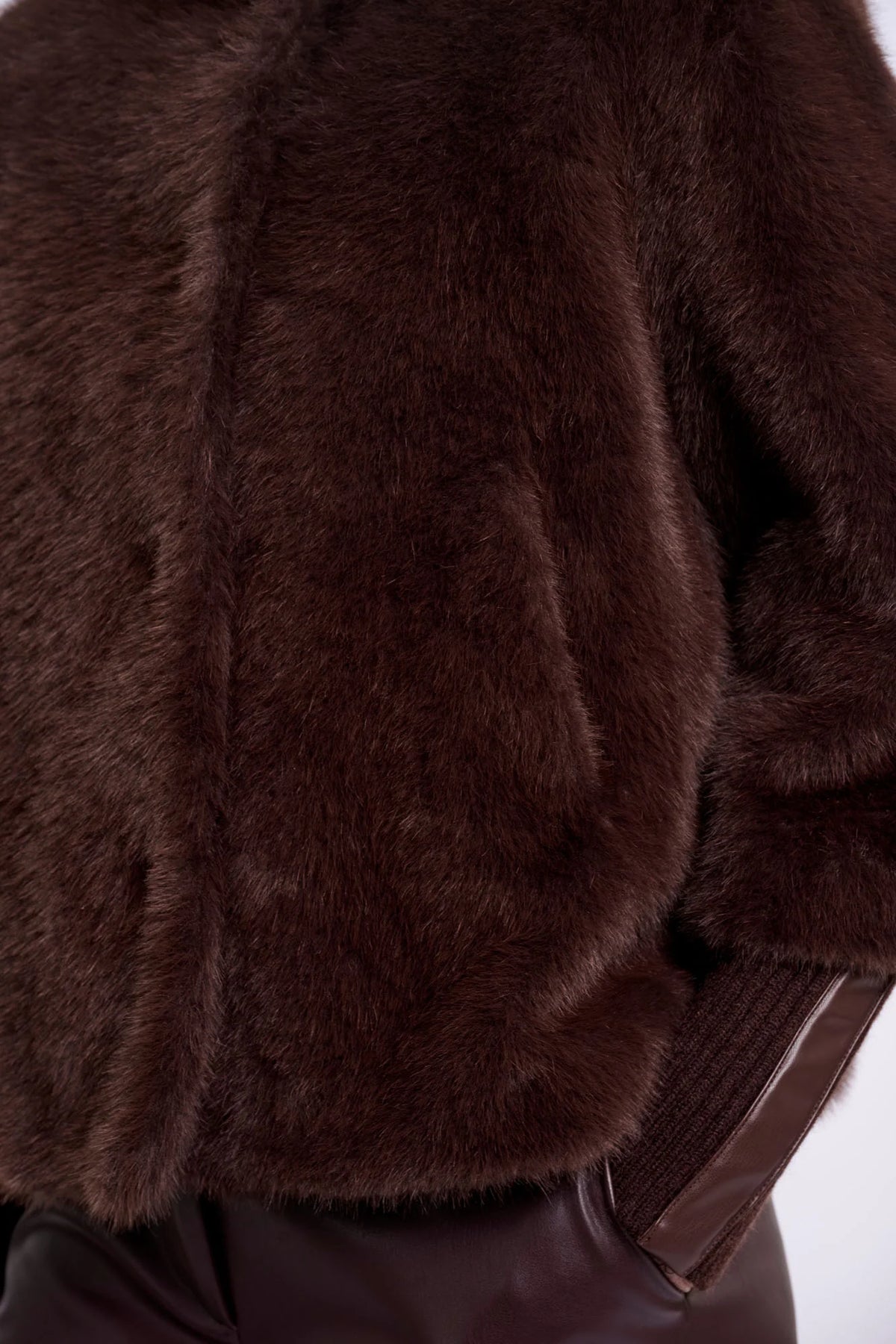 Close-up of a dark brown faux fur jacket with a ribbed brown knit sleeve, shown on a model wearing dark brown pants against a white background.