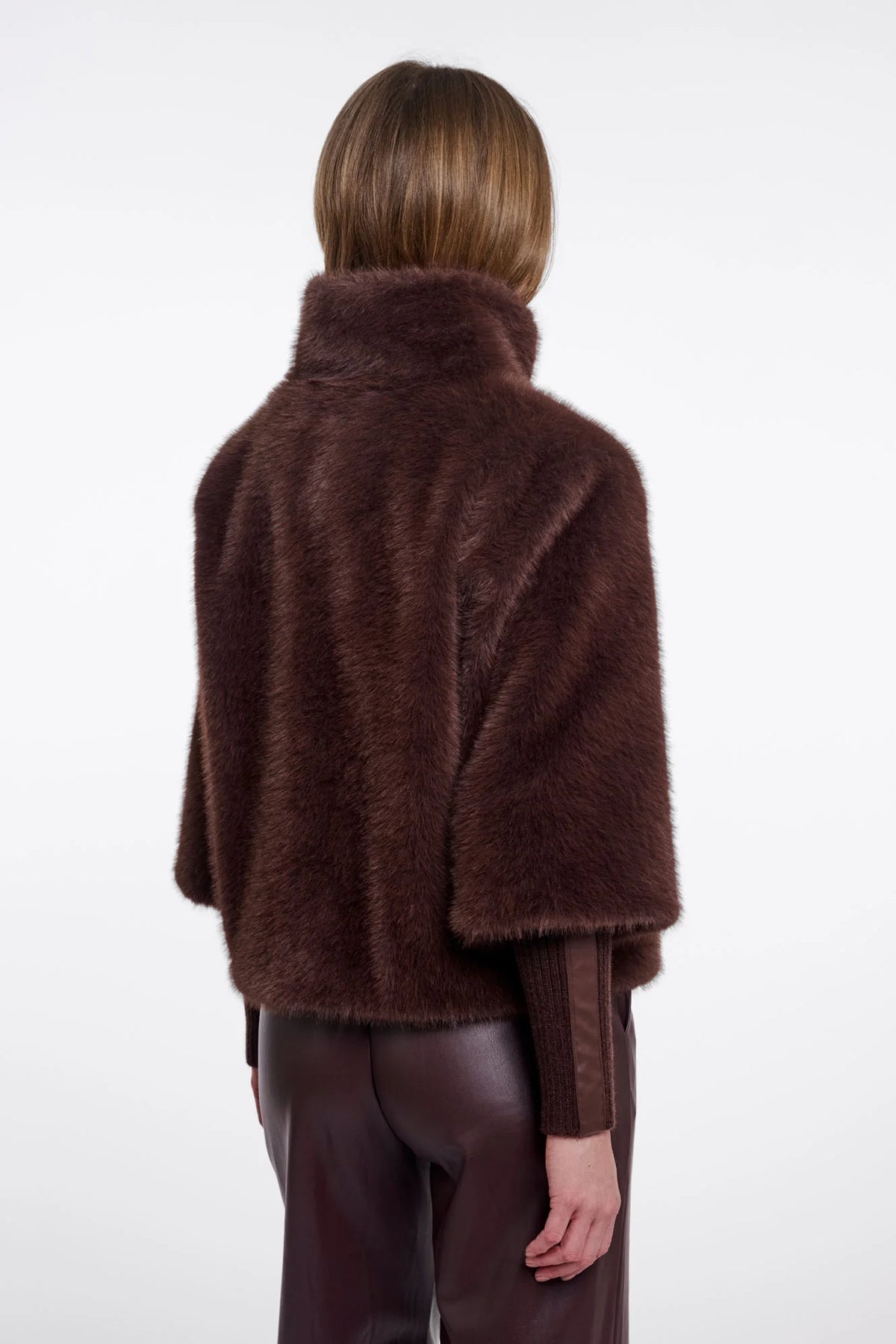 A model shown from the back wearing a dark brown faux fur cape jacket over a matching brown top and dark brown pants, set against a plain white studio background.
