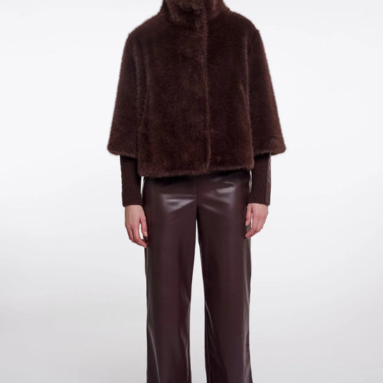 A model stands against a light gray studio background wearing a dark brown faux-fur cape jacket with a high collar, paired with matching dark brown wide-leg faux-leather pants and black pointed shoes.