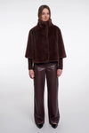 A model stands against a light gray studio background wearing a dark brown faux-fur cape jacket with a high collar, paired with matching dark brown wide-leg faux-leather pants and black pointed shoes.