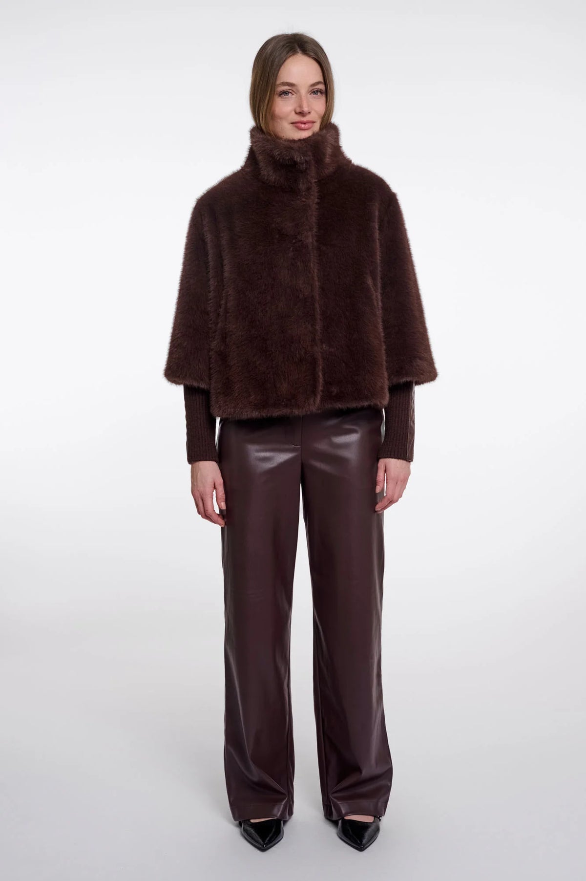 A model stands against a light gray studio background wearing a dark brown faux-fur cape jacket with a high collar, paired with matching dark brown wide-leg faux-leather pants and black pointed shoes.