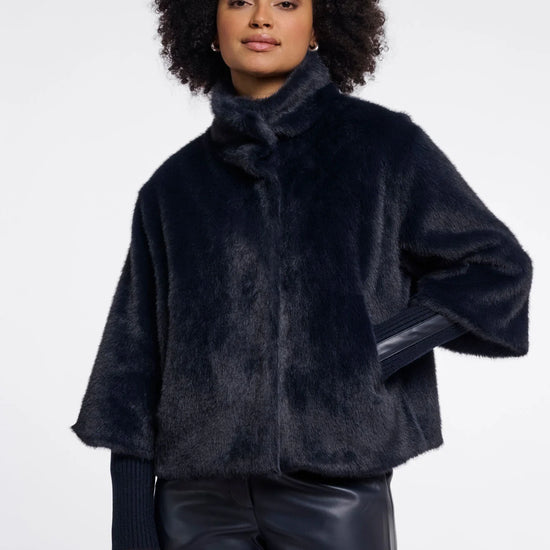 A model with curly dark hair wearing a dark navy blue faux fur jacket with a high collar and wide sleeves, paired with black pants, against a plain light gray background.