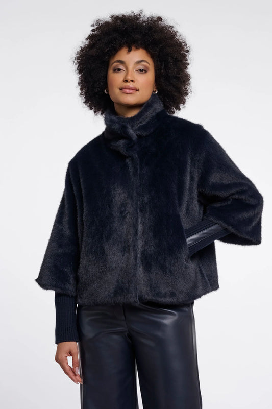 A model with curly dark hair wearing a dark navy blue faux fur jacket with a high collar and wide sleeves, paired with black pants, against a plain light gray background.