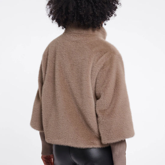 A person shown from the back wearing a cropped, fuzzy taupe-brown shrug or cardigan over a ribbed long-sleeve top and dark pants, against a plain light gray studio background.