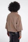 A person shown from the back wearing a cropped, fuzzy taupe-brown shrug or cardigan over a ribbed long-sleeve top and dark pants, against a plain light gray studio background.