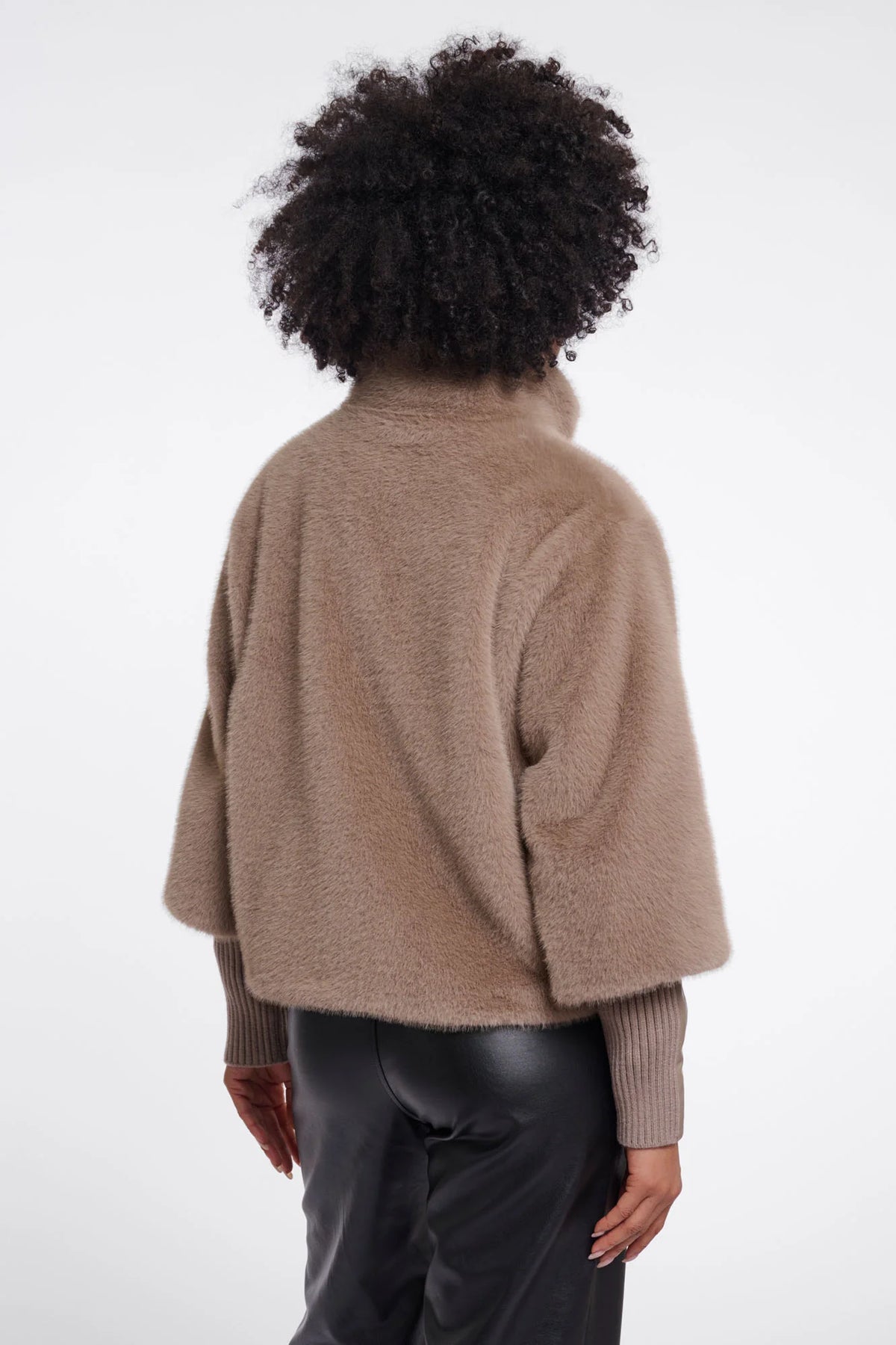 A person shown from the back wearing a cropped, fuzzy taupe-brown shrug or cardigan over a ribbed long-sleeve top and dark pants, against a plain light gray studio background.