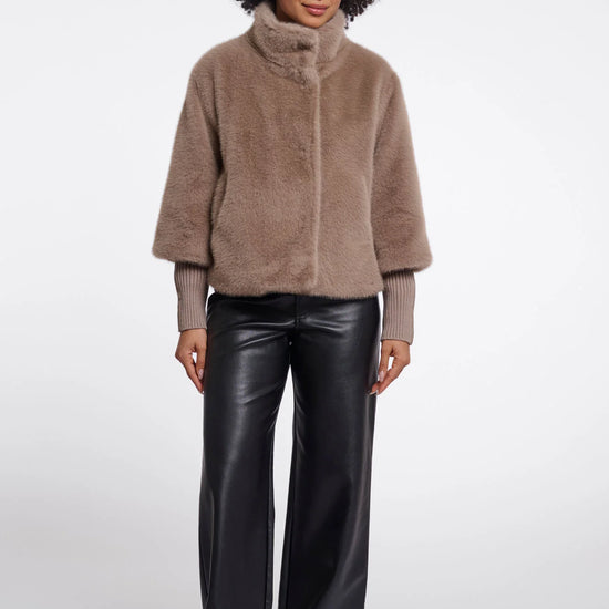 A model with an afro hairstyle wearing a taupe-brown fuzzy cropped jacket with a high mock neck and black wide-leg faux leather pants, standing against a light gray studio background.