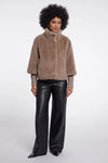 A model with an afro hairstyle wearing a taupe-brown fuzzy cropped jacket with a high mock neck and black wide-leg faux leather pants, standing against a light gray studio background.