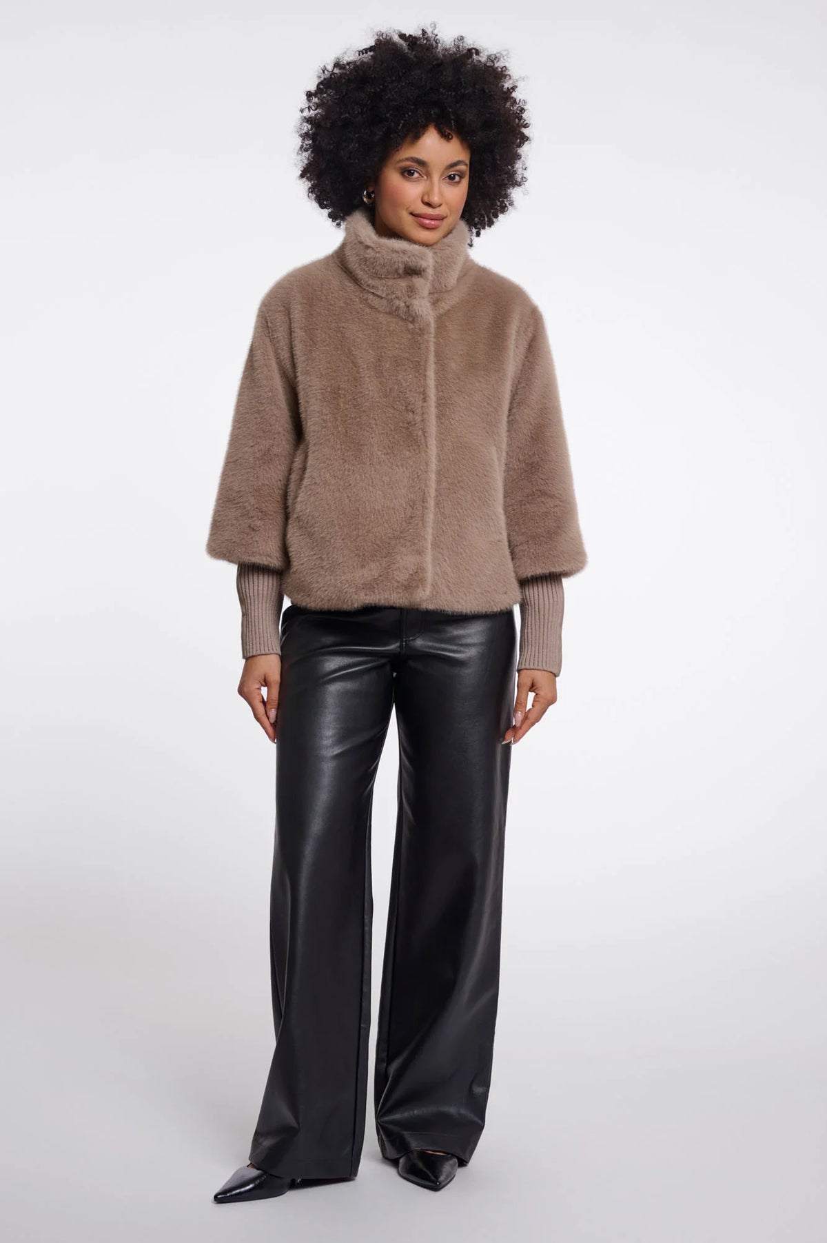 A model with an afro hairstyle wearing a taupe-brown fuzzy cropped jacket with a high mock neck and black wide-leg faux leather pants, standing against a light gray studio background.