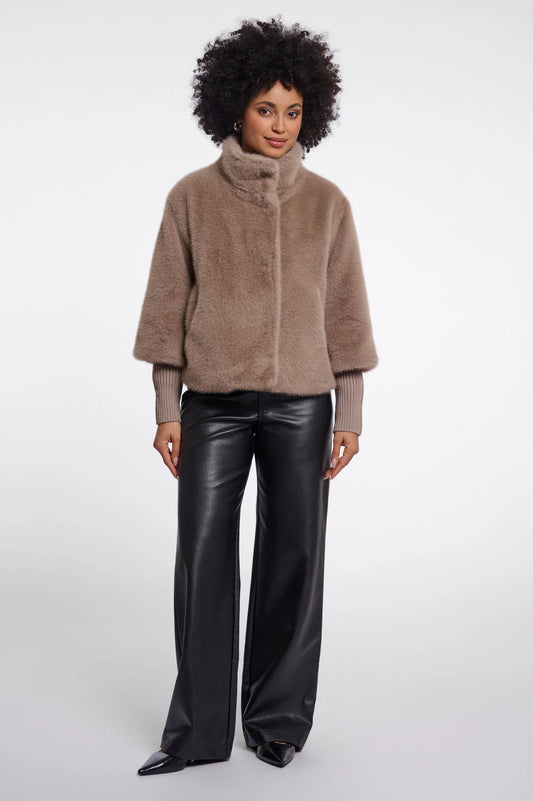A model with an afro hairstyle wearing a taupe-brown fuzzy cropped jacket with a high mock neck and black wide-leg faux leather pants, standing against a light gray studio background.