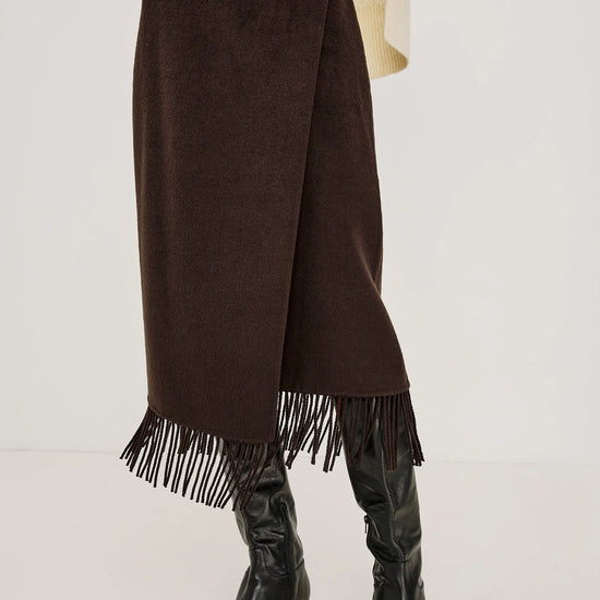 A dark brown wrap-style midi skirt with fringe trim at the hem, shown on a model wearing a cream sweater and black knee-high boots against a plain light background.