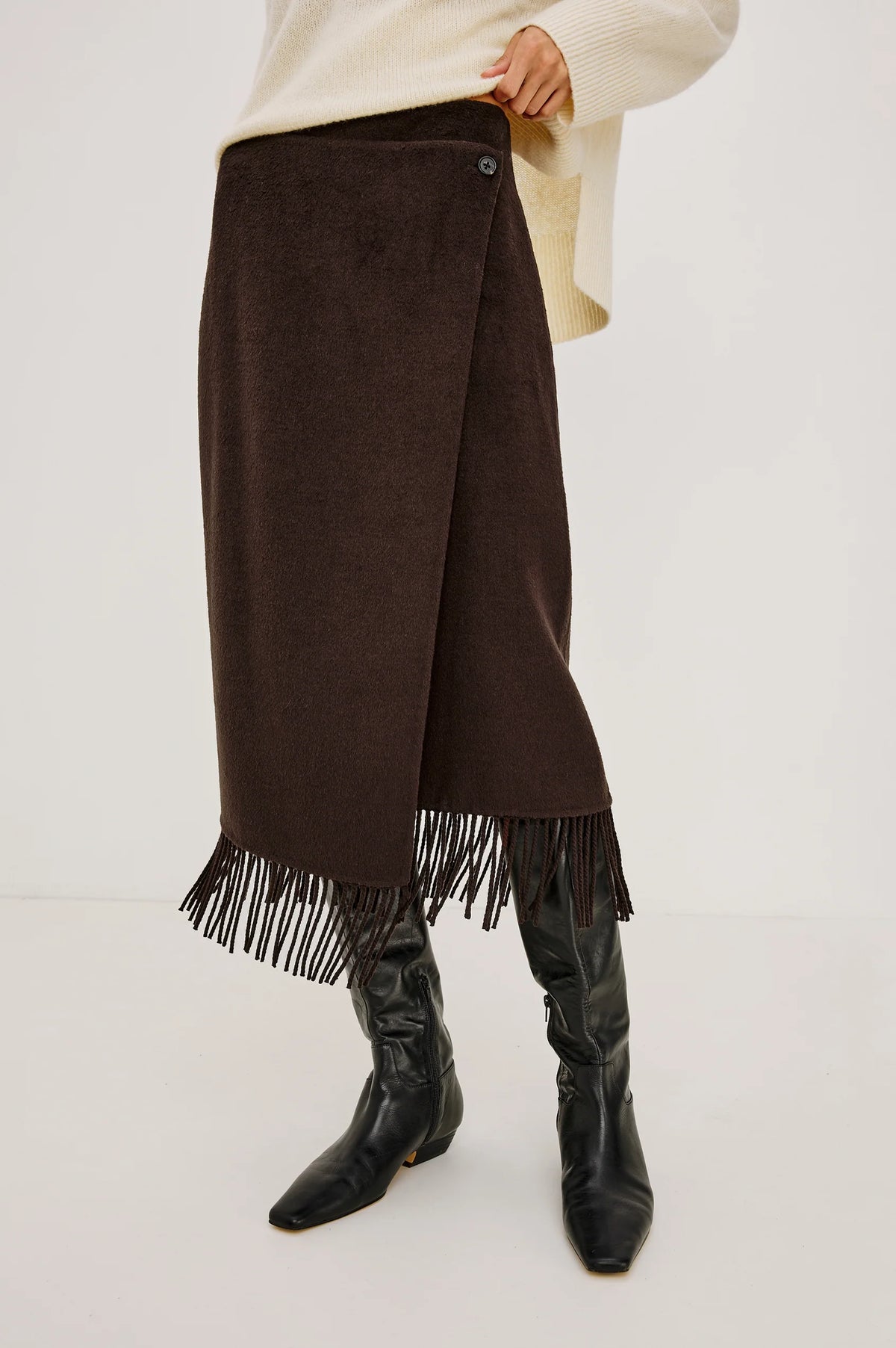 A dark brown wrap-style midi skirt with fringe trim at the hem, shown on a model wearing a cream sweater and black knee-high boots against a plain light background.