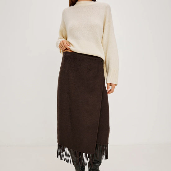 A model stands against a plain light gray studio background wearing a cream knit sweater, a dark brown midi skirt with fringe, and black pointed boots.