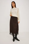 A model stands against a plain light gray studio background wearing a cream knit sweater, a dark brown midi skirt with fringe, and black pointed boots.