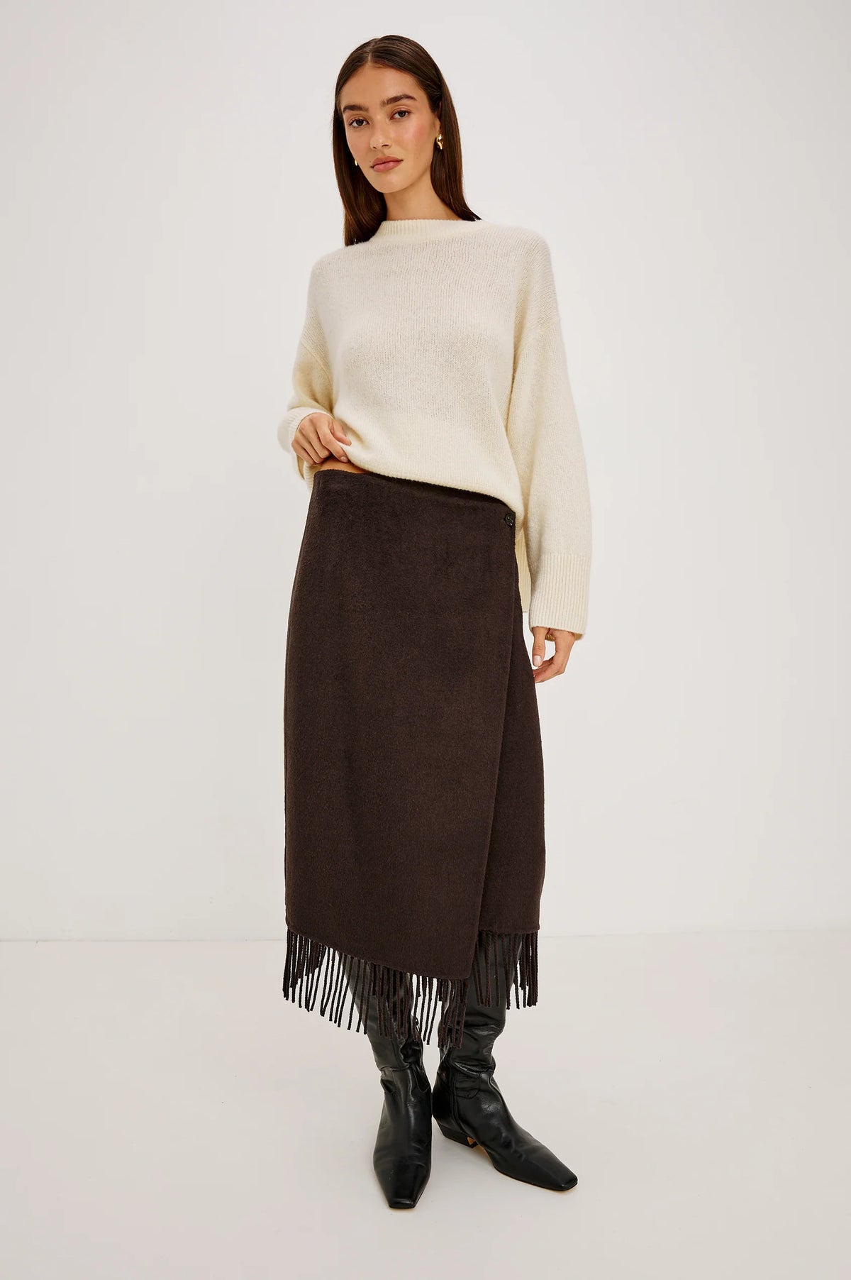 A model stands against a plain light gray studio background wearing a cream knit sweater, a dark brown midi skirt with fringe, and black pointed boots.