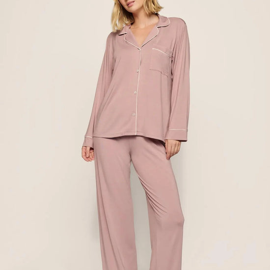 A woman modeling a two-piece dusty rose pajama set with white piping details, consisting of a long-sleeve button-up shirt and matching wide-leg pants, against a neutral studio backdrop.