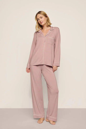 A woman modeling a two-piece dusty rose pajama set with white piping details, consisting of a long-sleeve button-up shirt and matching wide-leg pants, against a neutral studio backdrop.