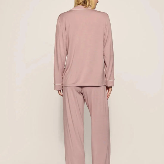 A woman viewed from the back wearing a dusty pink pajama set with long-sleeve top and wide-leg pants, standing against a plain light beige studio background.