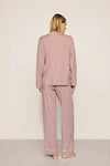 A woman viewed from the back wearing a dusty pink pajama set with long-sleeve top and wide-leg pants, standing against a plain light beige studio background.