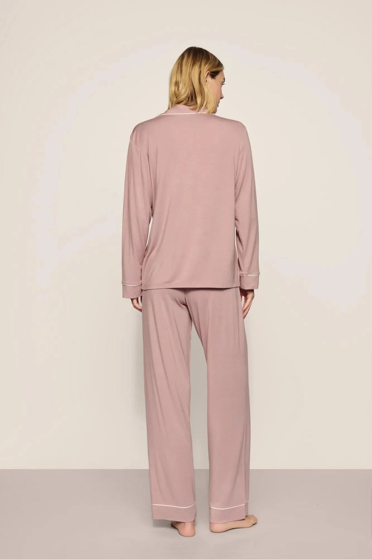 A woman viewed from the back wearing a dusty pink pajama set with long-sleeve top and wide-leg pants, standing against a plain light beige studio background.