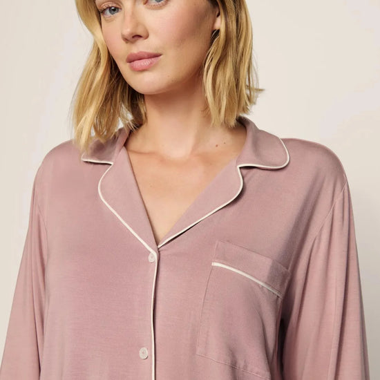 A woman wearing a dusty rose long-sleeve pajama shirt with white piping trim, shown from the chest up against a plain light beige background.