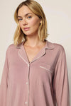 A woman wearing a dusty rose long-sleeve pajama shirt with white piping trim, shown from the chest up against a plain light beige background.