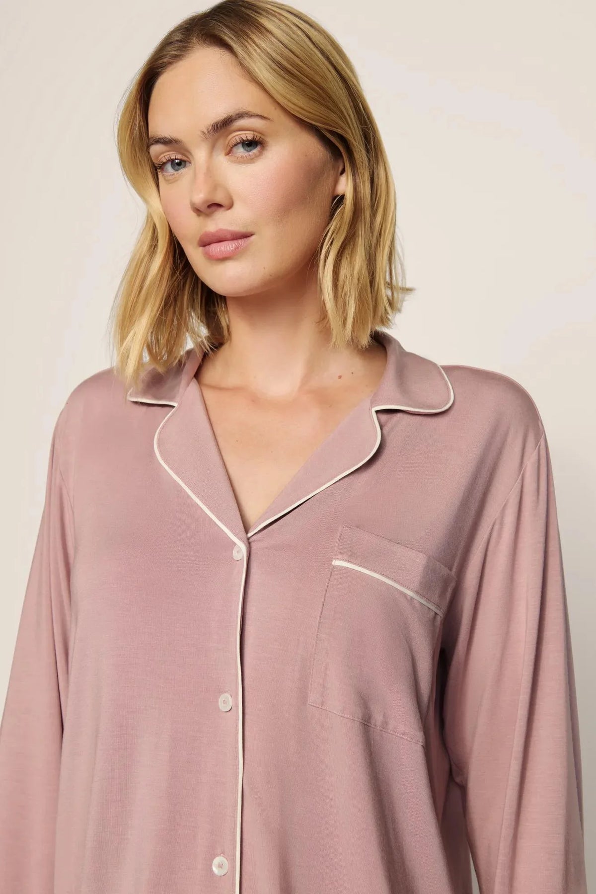 A woman wearing a dusty rose long-sleeve pajama shirt with white piping trim, shown from the chest up against a plain light beige background.