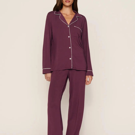 A woman modeling a two-piece pajama set in deep plum purple with white piping trim, standing against a plain light beige studio background.
