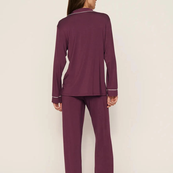 A woman stands barefoot against a plain light beige studio background, viewed from the back, wearing a matching long-sleeve pajama set in deep plum purple with thin white piping trim along the collar, cuffs, and hem.