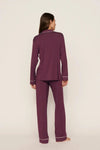 A woman stands barefoot against a plain light beige studio background, viewed from the back, wearing a matching long-sleeve pajama set in deep plum purple with thin white piping trim along the collar, cuffs, and hem.