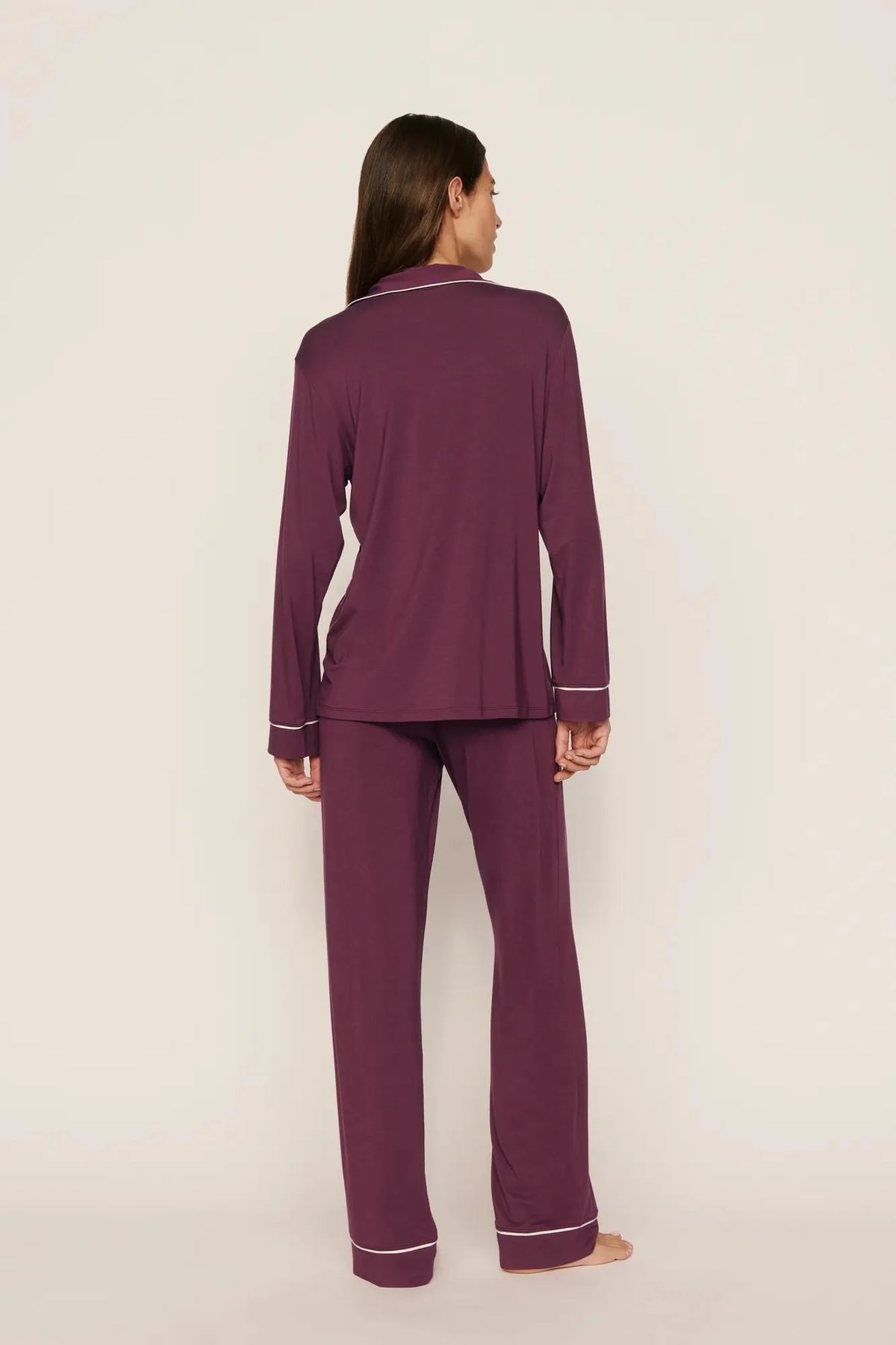 A woman stands barefoot against a plain light beige studio background, viewed from the back, wearing a matching long-sleeve pajama set in deep plum purple with thin white piping trim along the collar, cuffs, and hem.