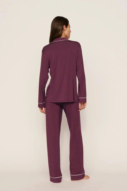 A woman stands barefoot against a plain light beige studio background, viewed from the back, wearing a matching long-sleeve pajama set in deep plum purple with thin white piping trim along the collar, cuffs, and hem.