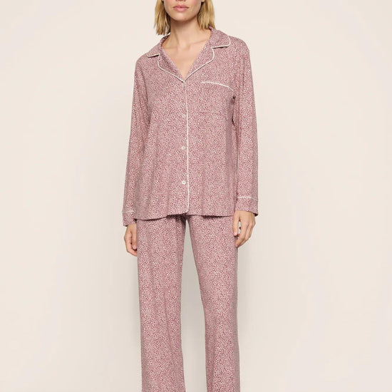 A woman modeling a two-piece pajama set with a pink and white micro-pattern. The set includes a long-sleeve button-up shirt with a collar and matching wide-leg pants, photographed against a neutral beige studio background.