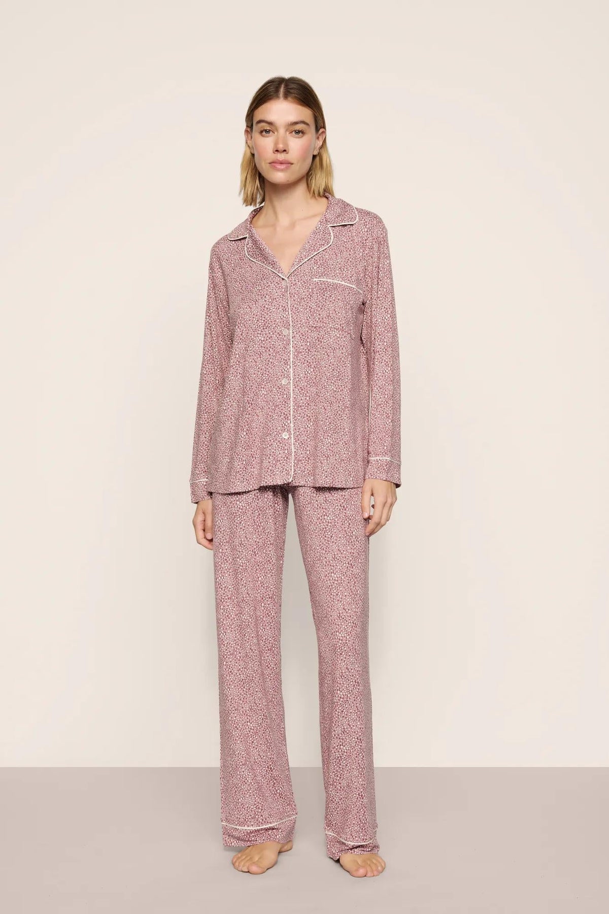 A woman modeling a two-piece pajama set with a pink and white micro-pattern. The set includes a long-sleeve button-up shirt with a collar and matching wide-leg pants, photographed against a neutral beige studio background.