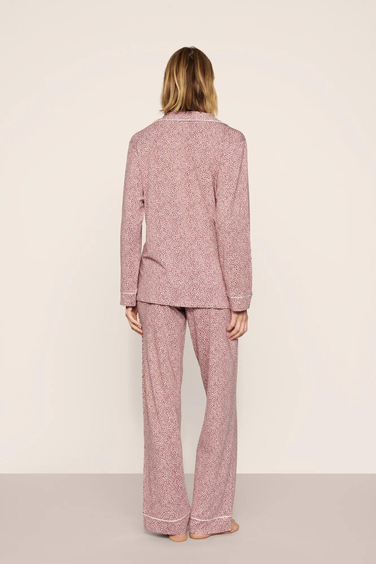 A woman viewed from behind wearing a matching two-piece pajama set with a textured, speckled pattern in dusty pink and white. The set includes a long-sleeve top and wide-leg pants, photographed against a neutral, light beige studio background.