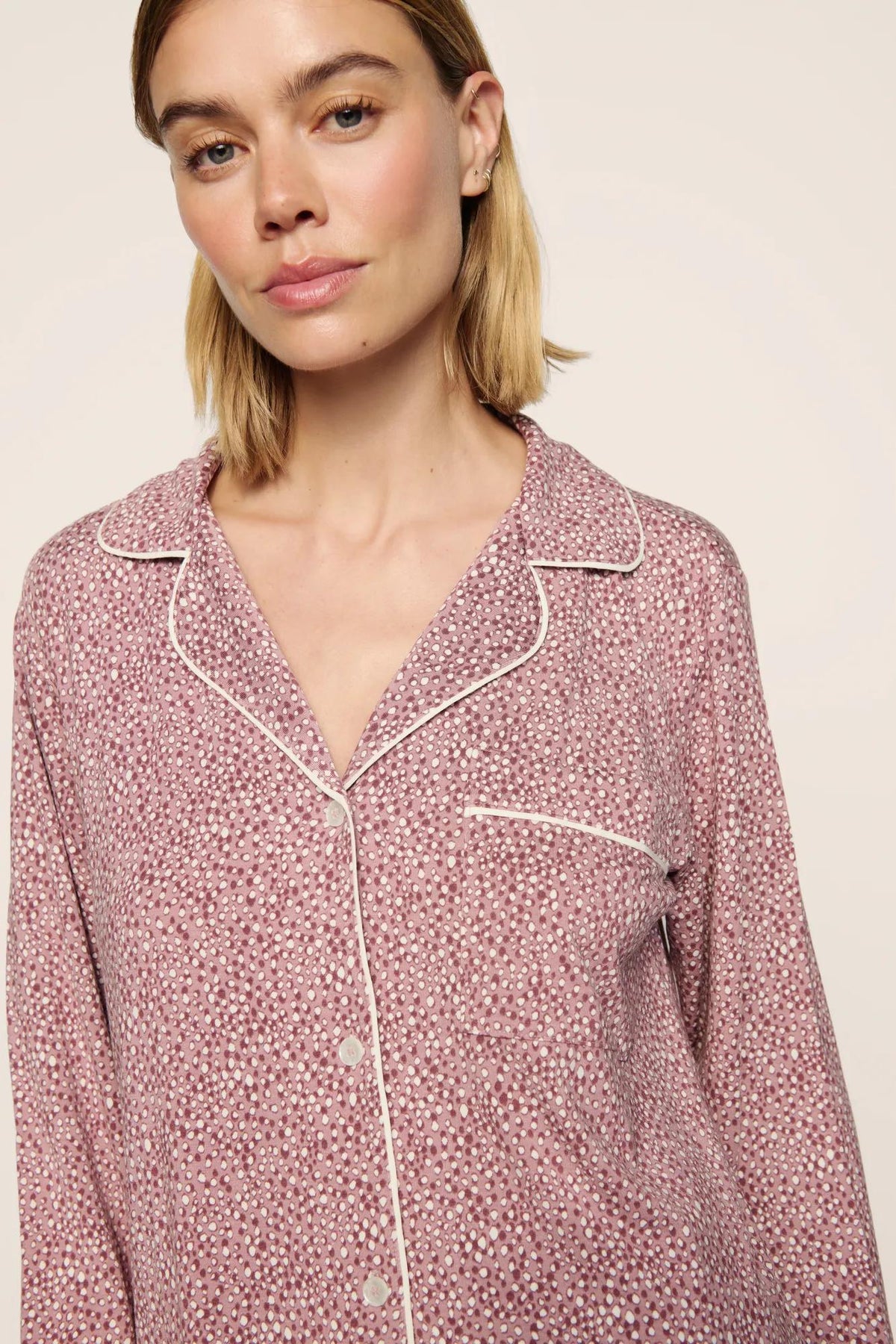 A close-up of a woman wearing a pink and white patterned long-sleeve pajama top with white piping details, set against a plain light beige background.
