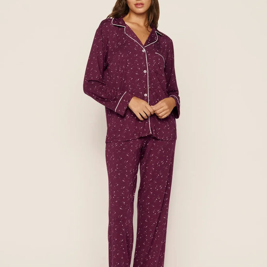 A woman modeling a matching two-piece pajama set in deep purple with a small white floral pattern and white piping details, standing against a plain off-white studio background.