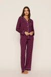 A woman modeling a matching two-piece pajama set in deep purple with a small white floral pattern and white piping details, standing against a plain off-white studio background.