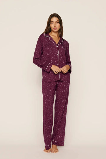 A woman modeling a matching two-piece pajama set in deep purple with a small white floral pattern and white piping details, standing against a plain off-white studio background.