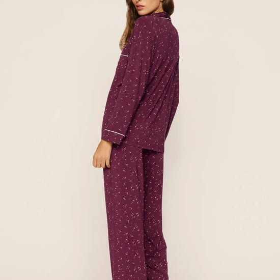 A woman modeling a two-piece pajama set in a deep purple color with a small white floral pattern and white piping details, shown from a side-back angle against a plain off-white studio background.