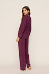 A woman modeling a two-piece pajama set in a deep purple color with a small white floral pattern and white piping details, shown from a side-back angle against a plain off-white studio background.