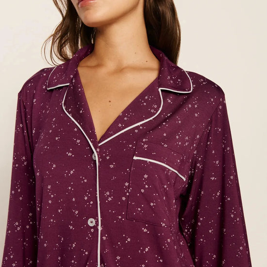 A woman wearing a long-sleeve button-front pajama shirt in deep purple with a small white floral print and white piping trim, shown against a plain light beige background.