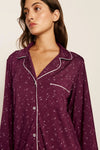 A woman wearing a long-sleeve button-front pajama shirt in deep purple with a small white floral print and white piping trim, shown against a plain light beige background.